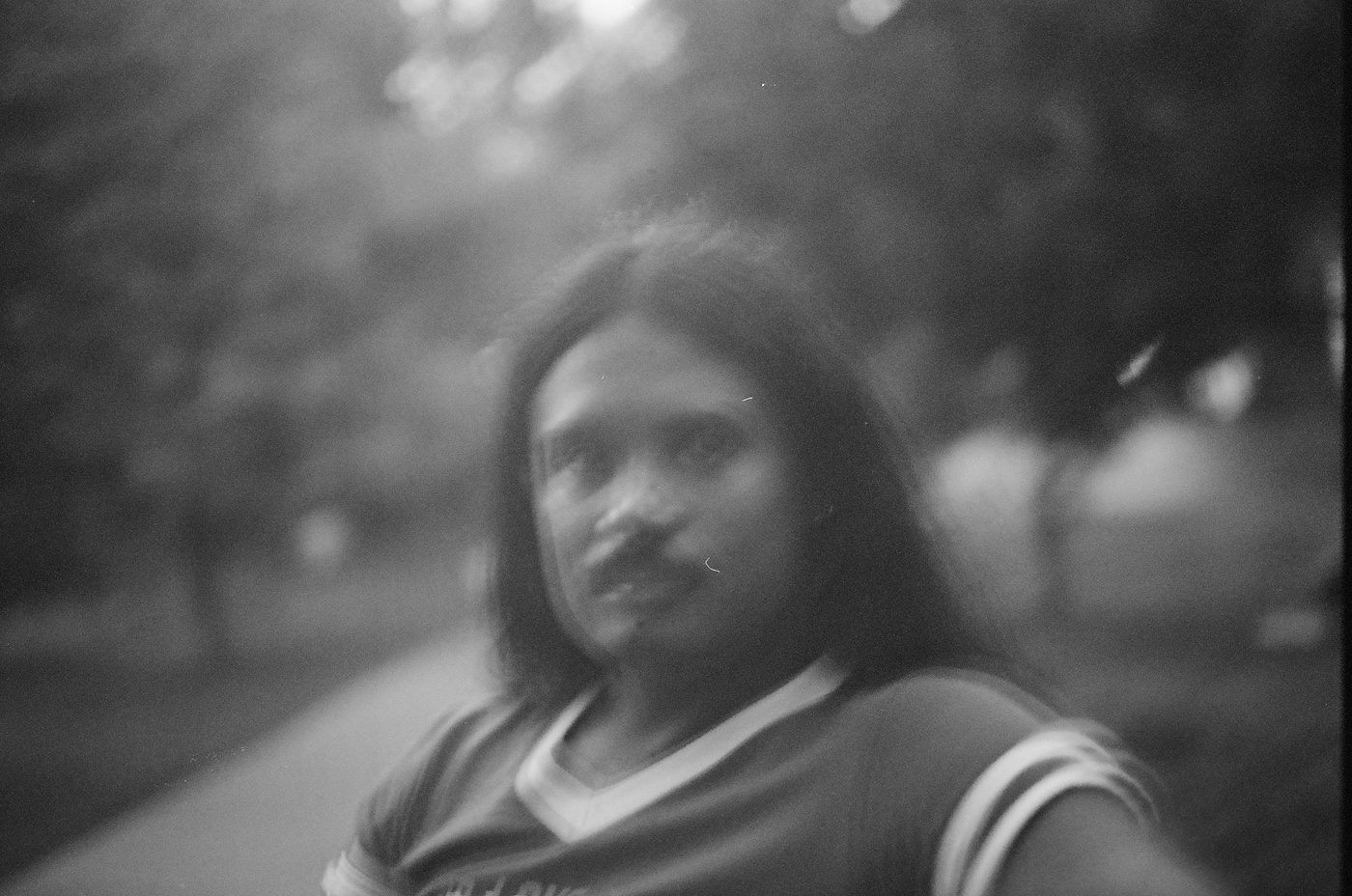 A black-and-white portrait of a person with long dark hair and a pale-collared shirt, looking to camera, the background thrown well out of focus.
