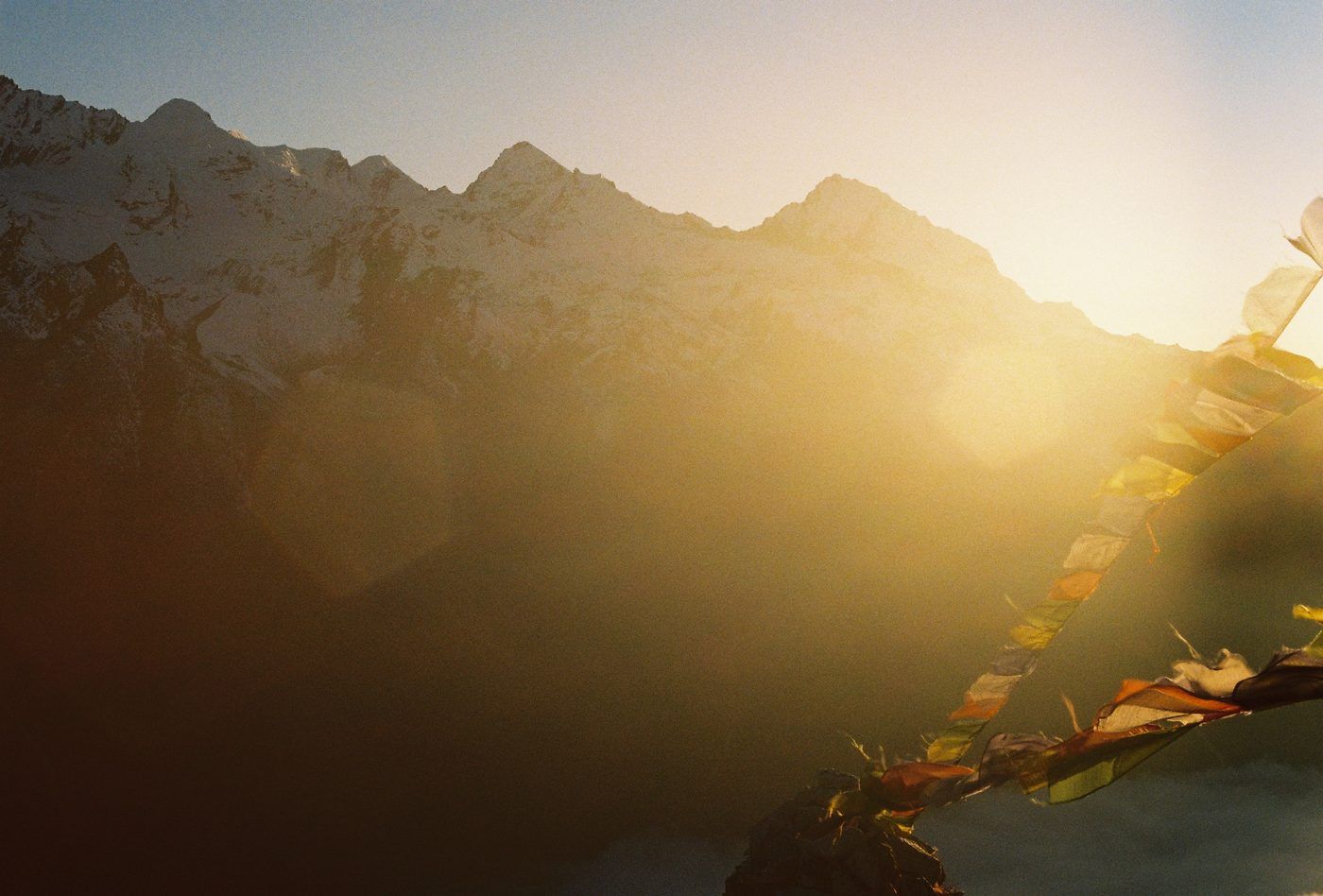 Mountain ridges in near-silhouette under an intense sun flare, with strings of coloured prayer flags crossing the lower right of the frame.